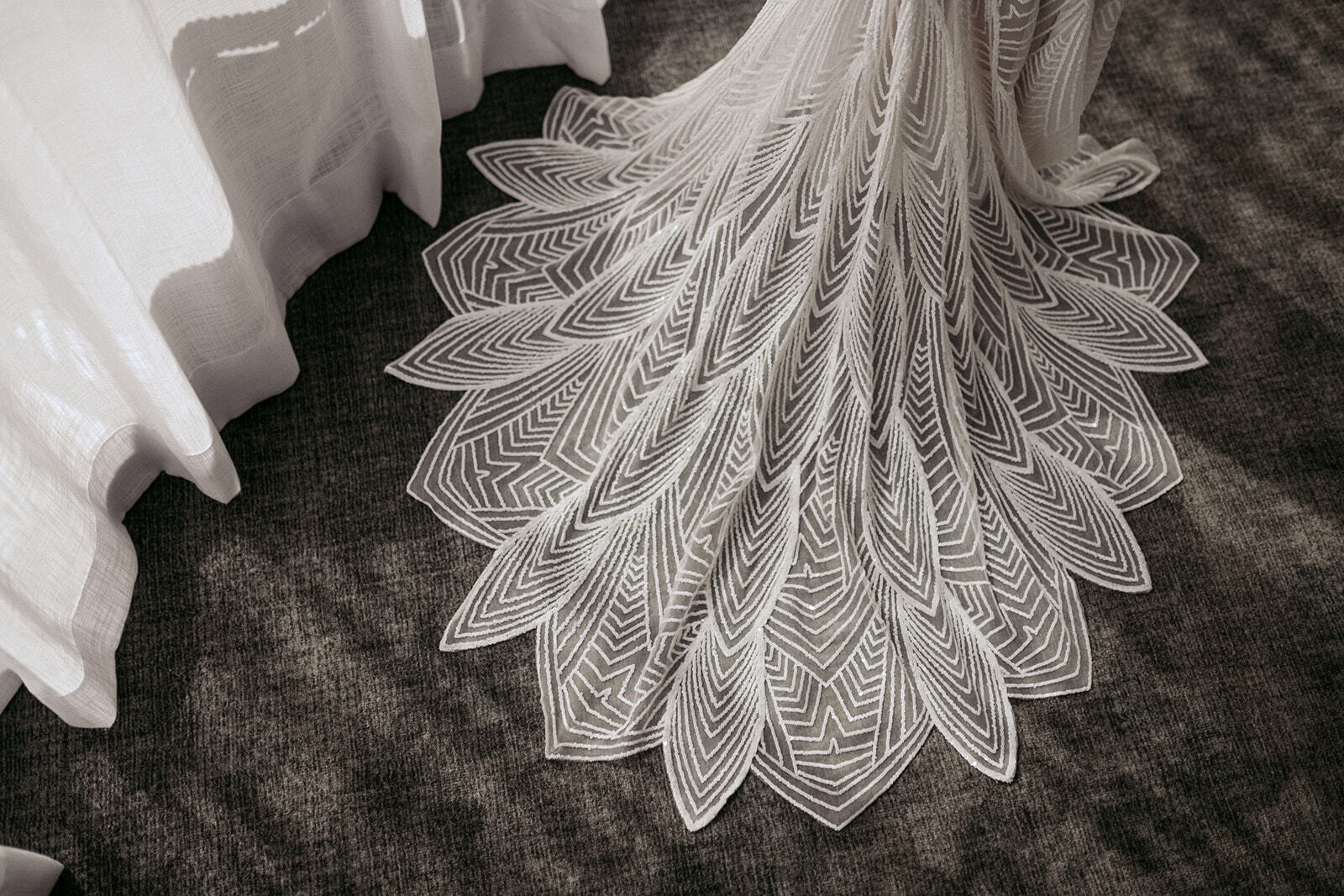 Close-up of an Alexis George wedding gown train featuring intricate geometric art deco lace patterns, displayed on a textured carpet in natural light.
