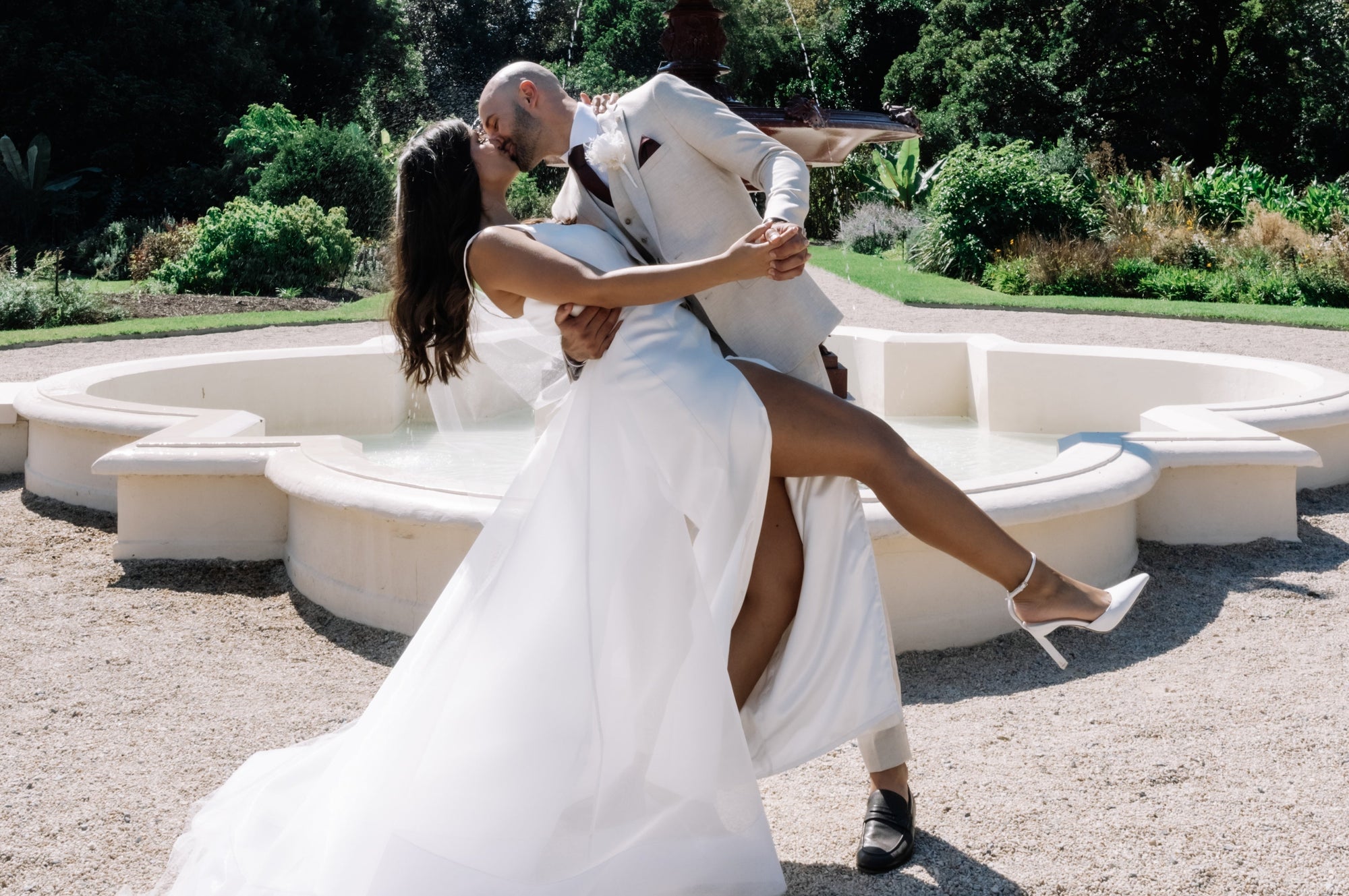 Groom dipping the bride in front of a decorative garden fountain at the Adelaide Botanic Gardens, the bride’s white wedding dress flowing and the couple sharing a joyful moment under a clear blue sky