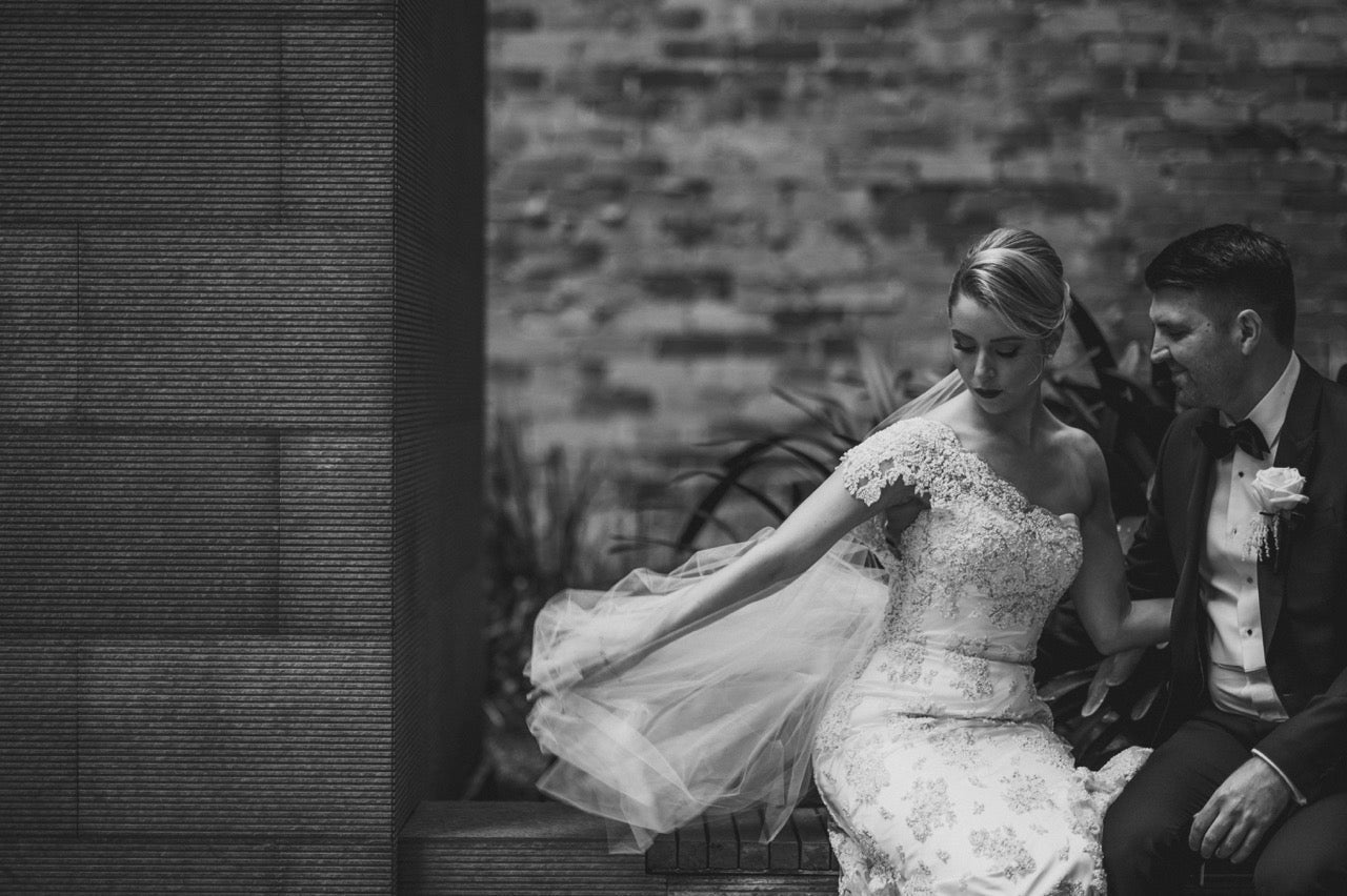 Bride and groom sitting together at their Adelaide wedding, bride wearing a bespoke Alexis George one-shoulder beaded lace gown with flowing veil, groom in a classic tuxedo, against a textured brick and stone backdrop.